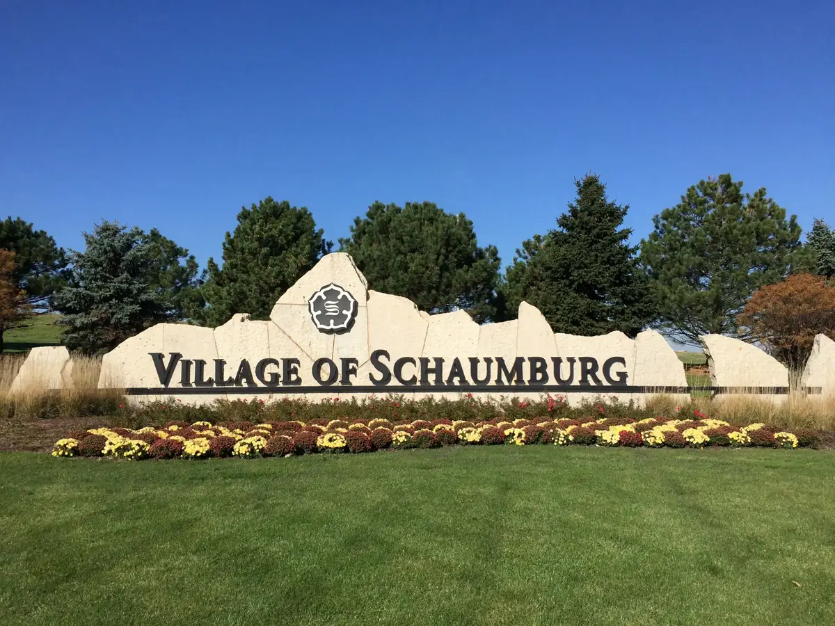 Village of Schaumburg welcome sign surrounded by flowers and greenery on a sunny day, representing Schaumburg, IL community.